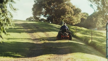 A man riding a mower in the middle of a green grass field with sunlight falling on it.