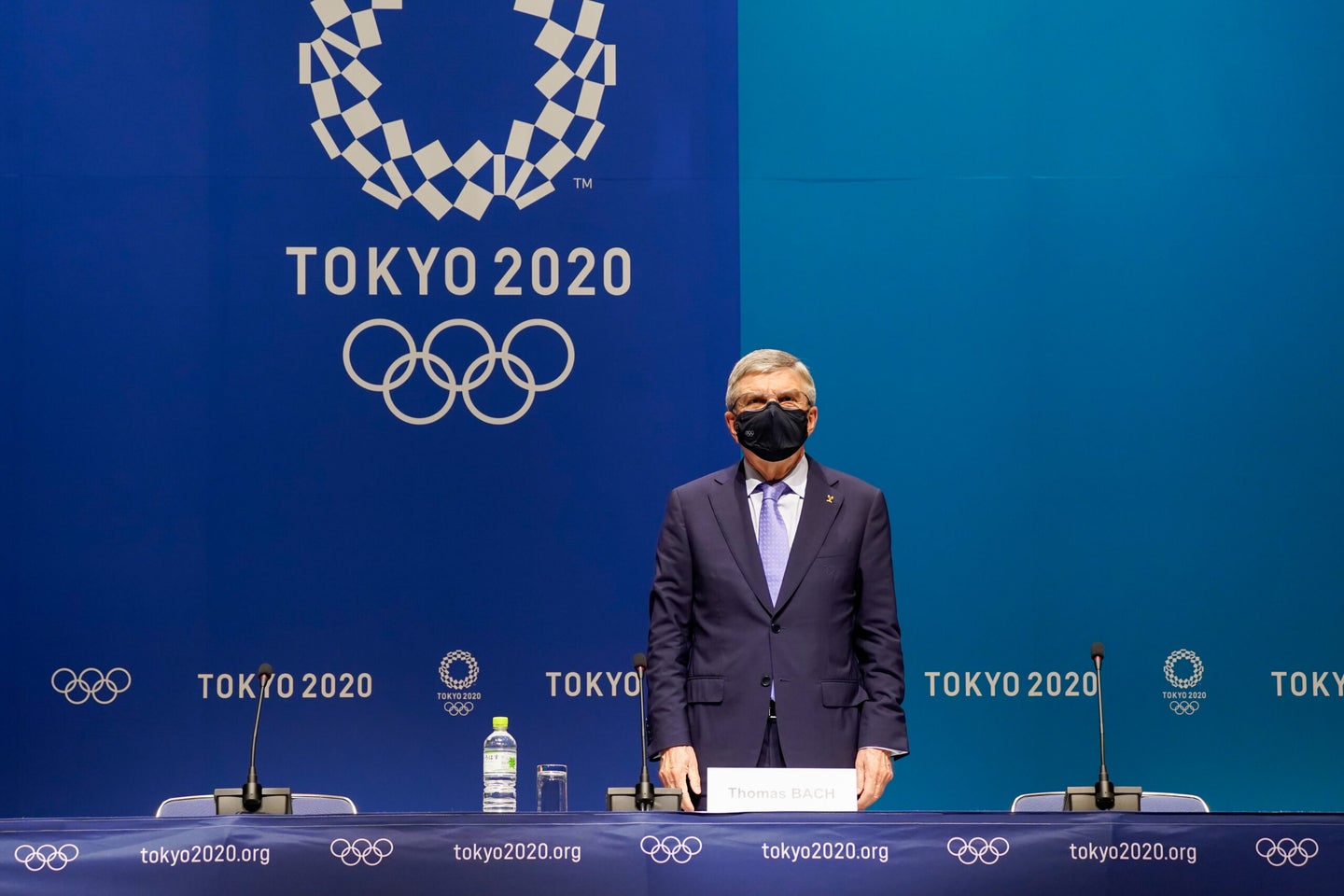 A grey haired man in a dark suit, wearing a black mask stands in from of a two-toned blue background and behind a long table set up with three microphones and chairs. The background says "Tokyo 2020" in large white letters and displays the Olympic logo.