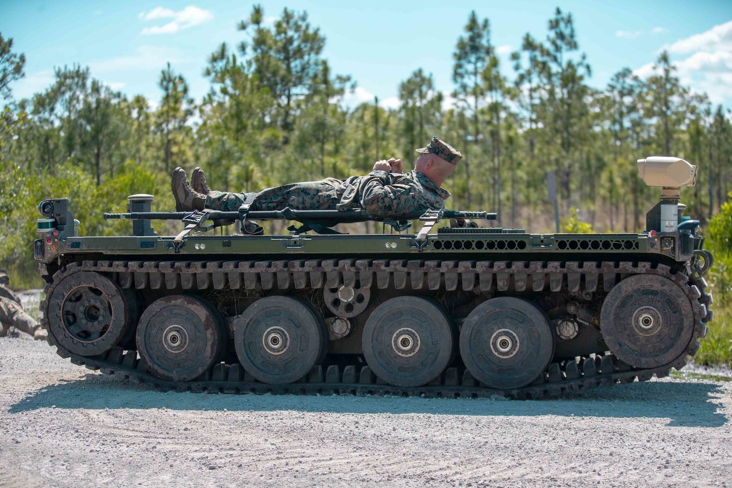 A Marine lies on top of a robotic vehicle on treads.