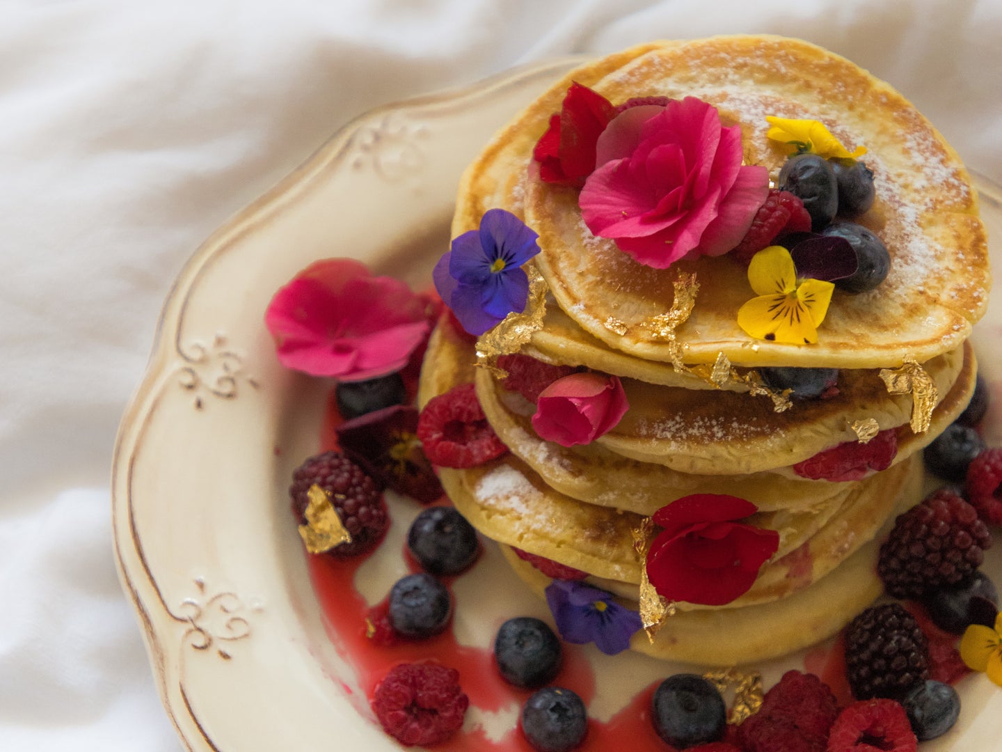 A stack of pancakes topped with berries and edible flowers, on a white plate atop a white tablecloth.