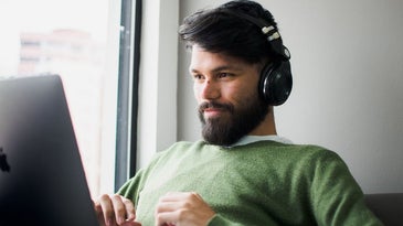 A young man with dark hair and beard and a pair of black headphones placed in his ear and a laptop in front of him.