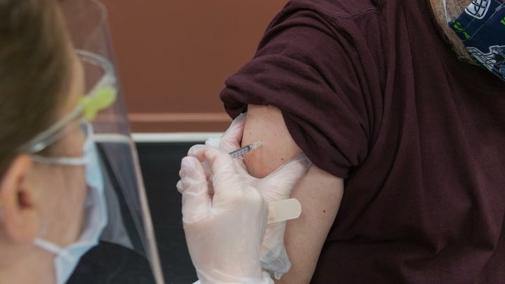 A healthcare professional administers a shot in an arm to a person in a brown shirt.