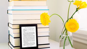 A Kobo e-reader leaning against a stack of books next to a glass jar with yellow flowers in it.
