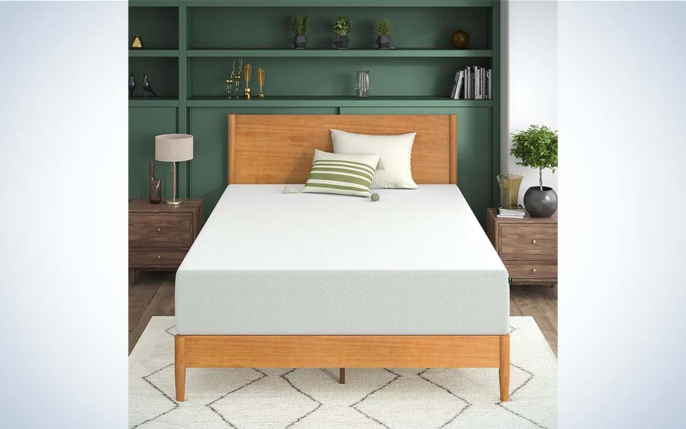 A white mattress made by Zinus in a wooden frame flanked by green shelves and a white rug.