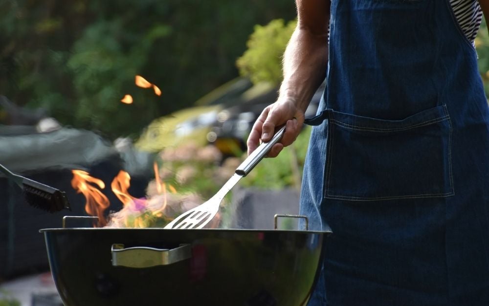 A man hand with a big silver fork for grilling into a black grill with a flame into it outside a yard.