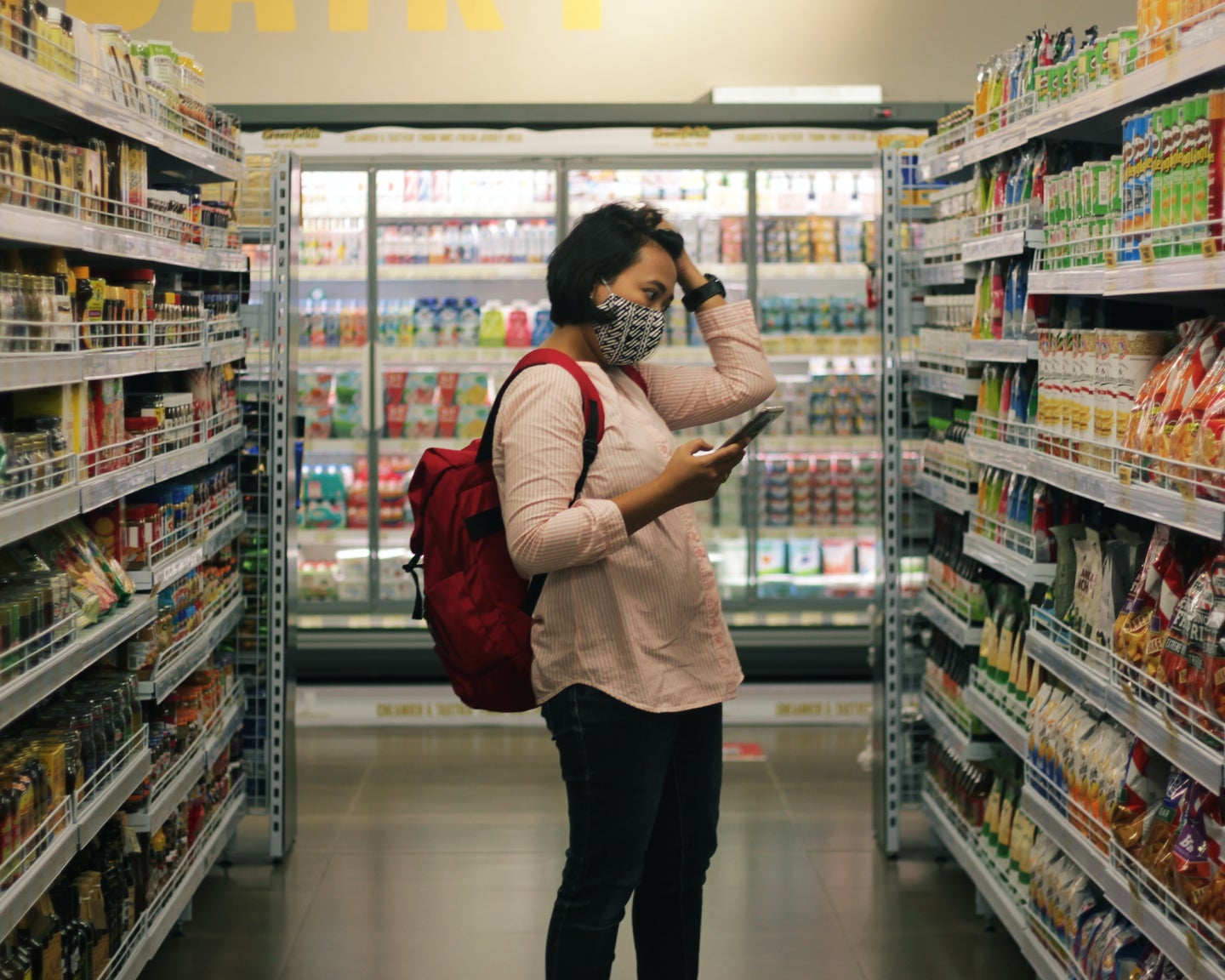 A dark haired person wearing a black and white patterned mask with a red backpack stands in a grocery store aisle, holding up a smartphone in one hand and running their other hand through their hair. They are looking intently at an array of colorful packaged foods.
