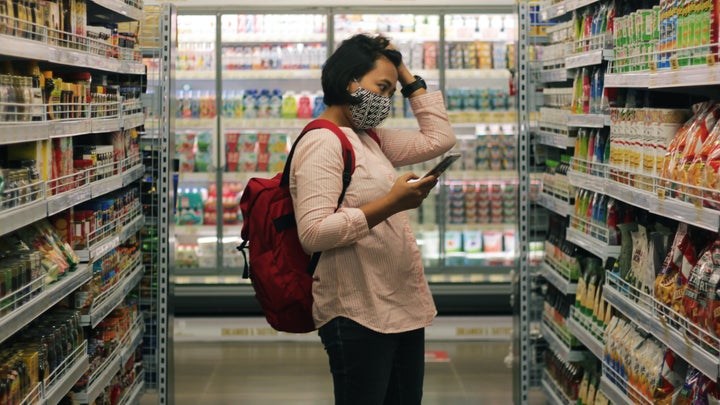 A dark haired person wearing a black and white patterned mask with a red backpack stands in a grocery store aisle, holding up a smartphone in one hand and running their other hand through their hair. They are looking intently at an array of colorful packaged foods.