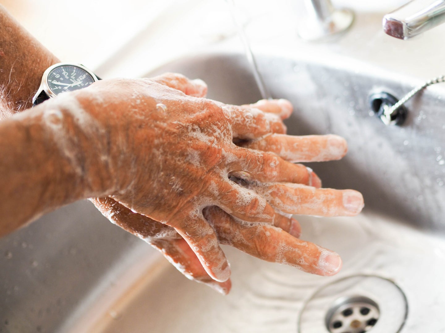 A person washing their hands with soap and water spreads soap between their fingers over the basin of a sink.