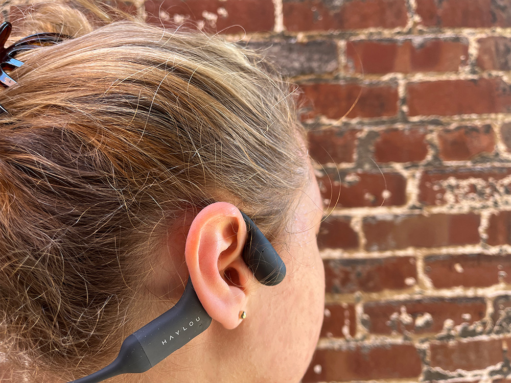A female-presenting person standing in front of a brick wall wearing a pair of grey Haylou bone conduction headphones