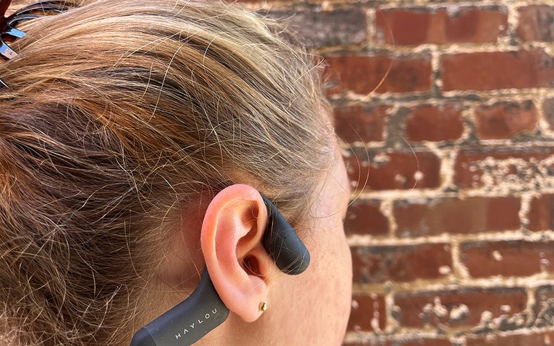 A female-presenting person standing in front of a brick wall wearing a pair of grey Haylou bone conduction headphones
