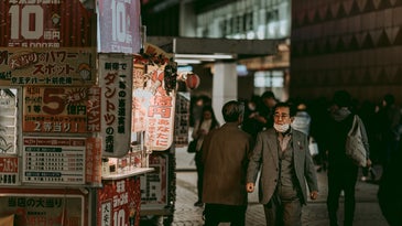 A man wearing a grey suit and glasses has a white surgical mask partially covering his mouth and chin. He is walking down a cobblestone street facing towards the camera while others walk facing the other way. It is nighttime and a building and street vendor are in the background.
