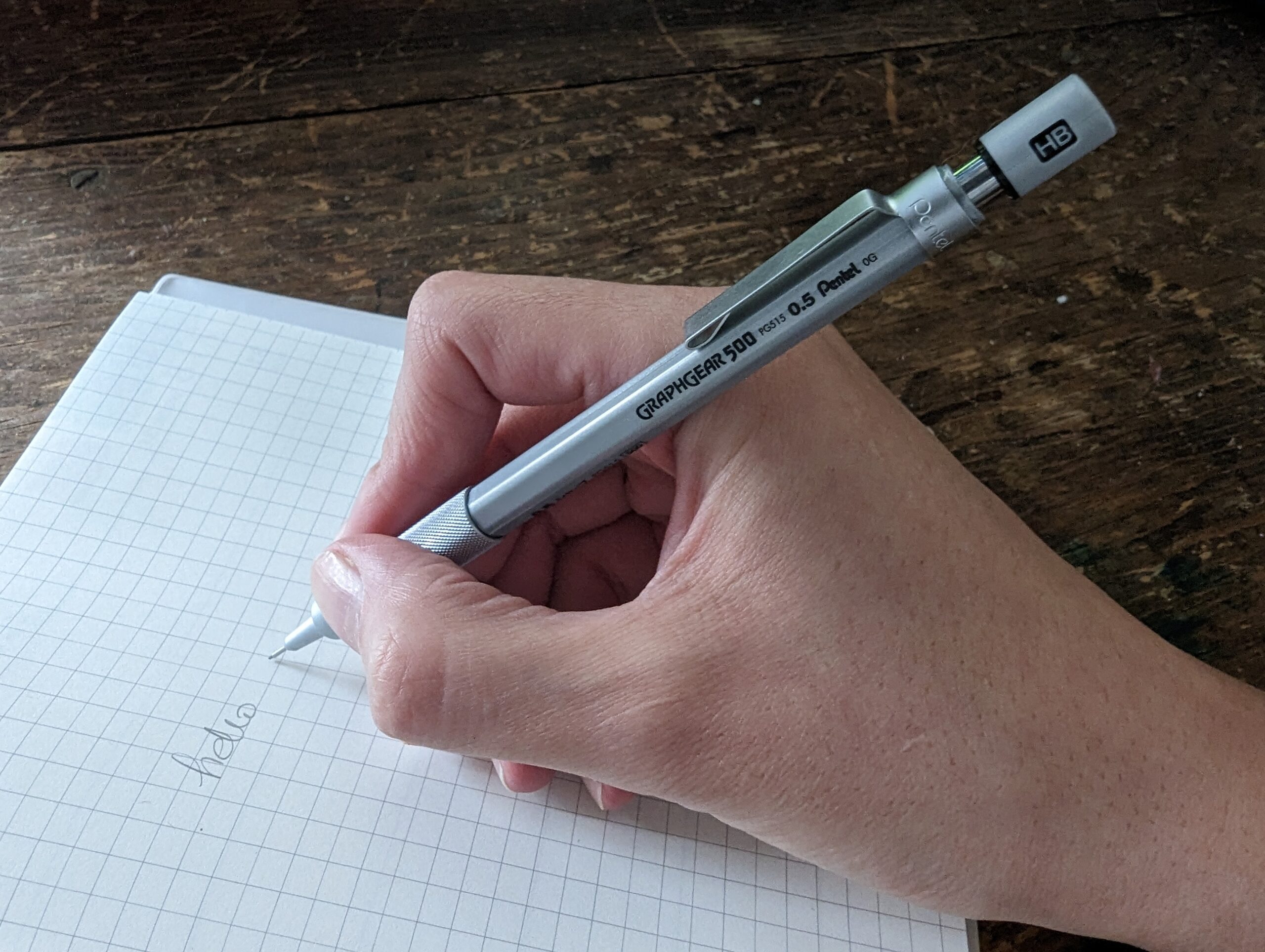 A person holding a mechanical pencil and writing "hello" on a piece of checkered notebook paper.
