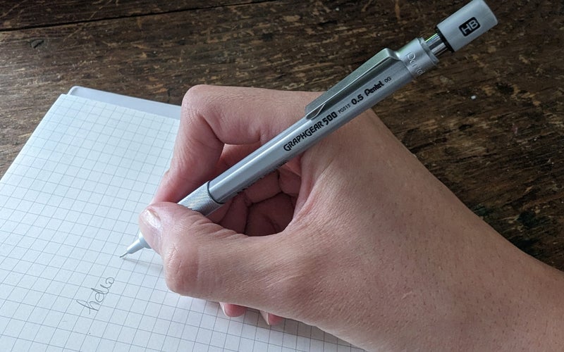 A person holding a mechanical pencil and writing "hello" on a piece of checkered notebook paper.