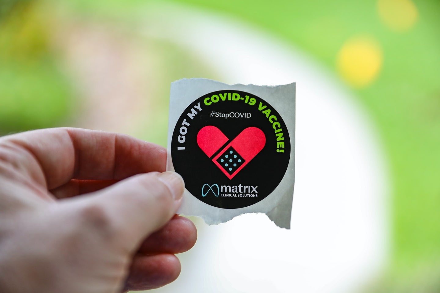 A hand holds a black sticker against a green background. The sticker has a red bandage on it and says 'I got my COVID-19 vaccine.'