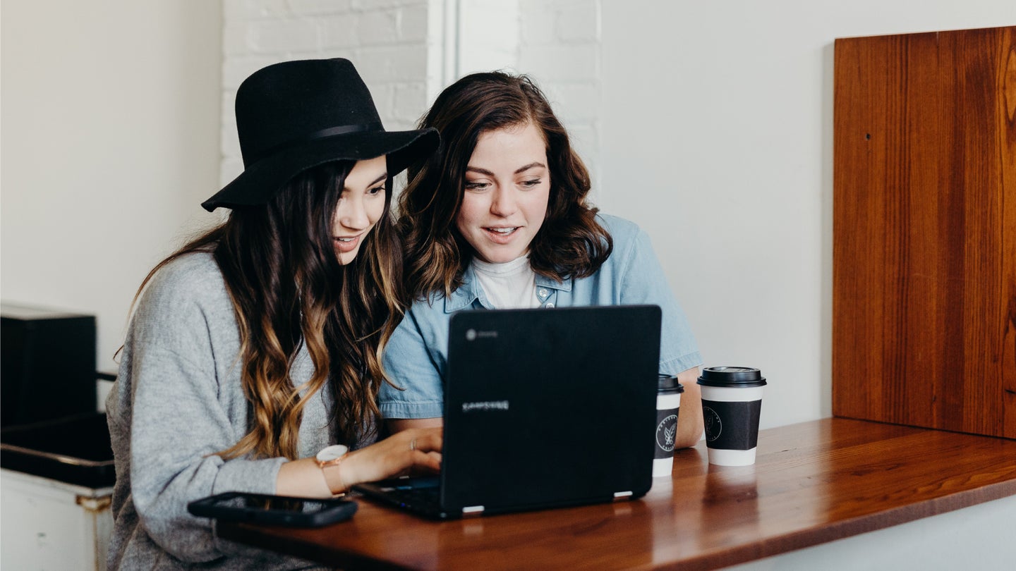 Two women using one laptop at a wooden table. One woman is wearing a black hat.
