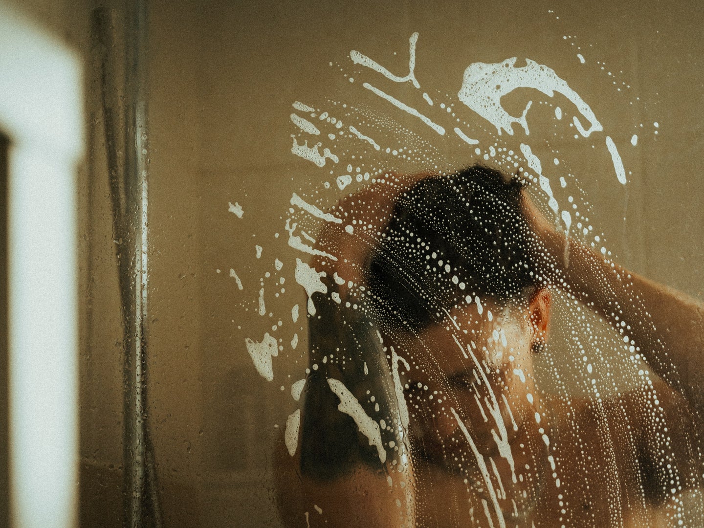 A person standing in a glass shower, washing their hair, with soap suds on the glass shower door.