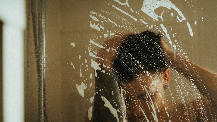 A person standing in a glass shower, washing their hair, with soap suds on the glass shower door.