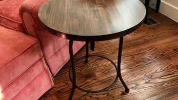 A refinished wooden side table with black iron legs, on a hardwood floor next to a pink armchair.