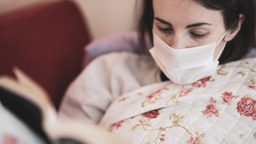 A woman in bed wears a mask and reads a book.