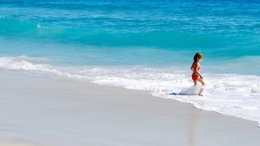 A young child standing alone at the edge of a bright blue ocean, playing in the foam.