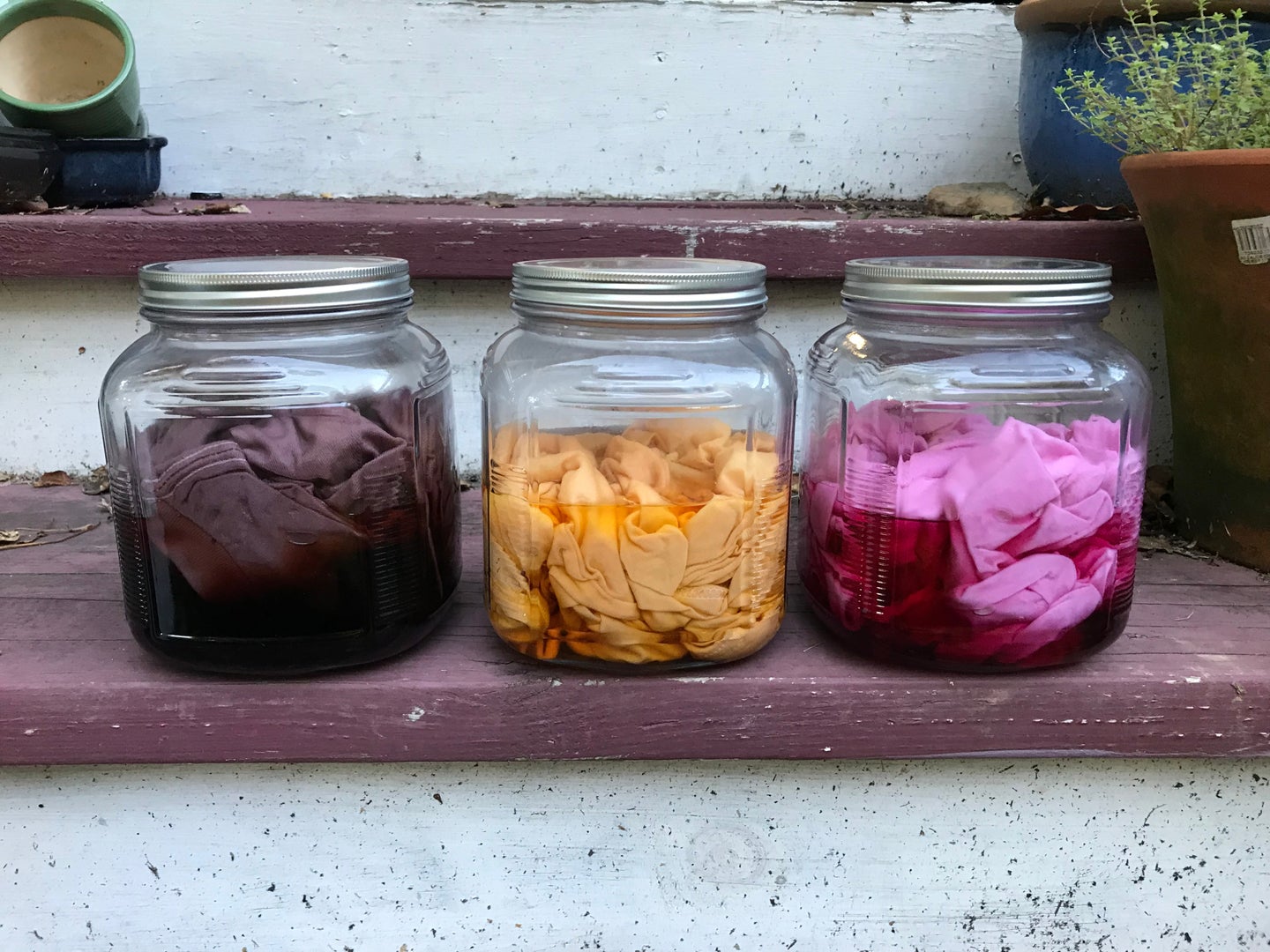 Three mason jars with homemade fabric dye and some dyed shirts inside them. From left to right: purple, orange, and pink.