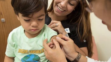 A small child is having a bandage put on his arm after receiving a shot. A caregiver stands behind him.