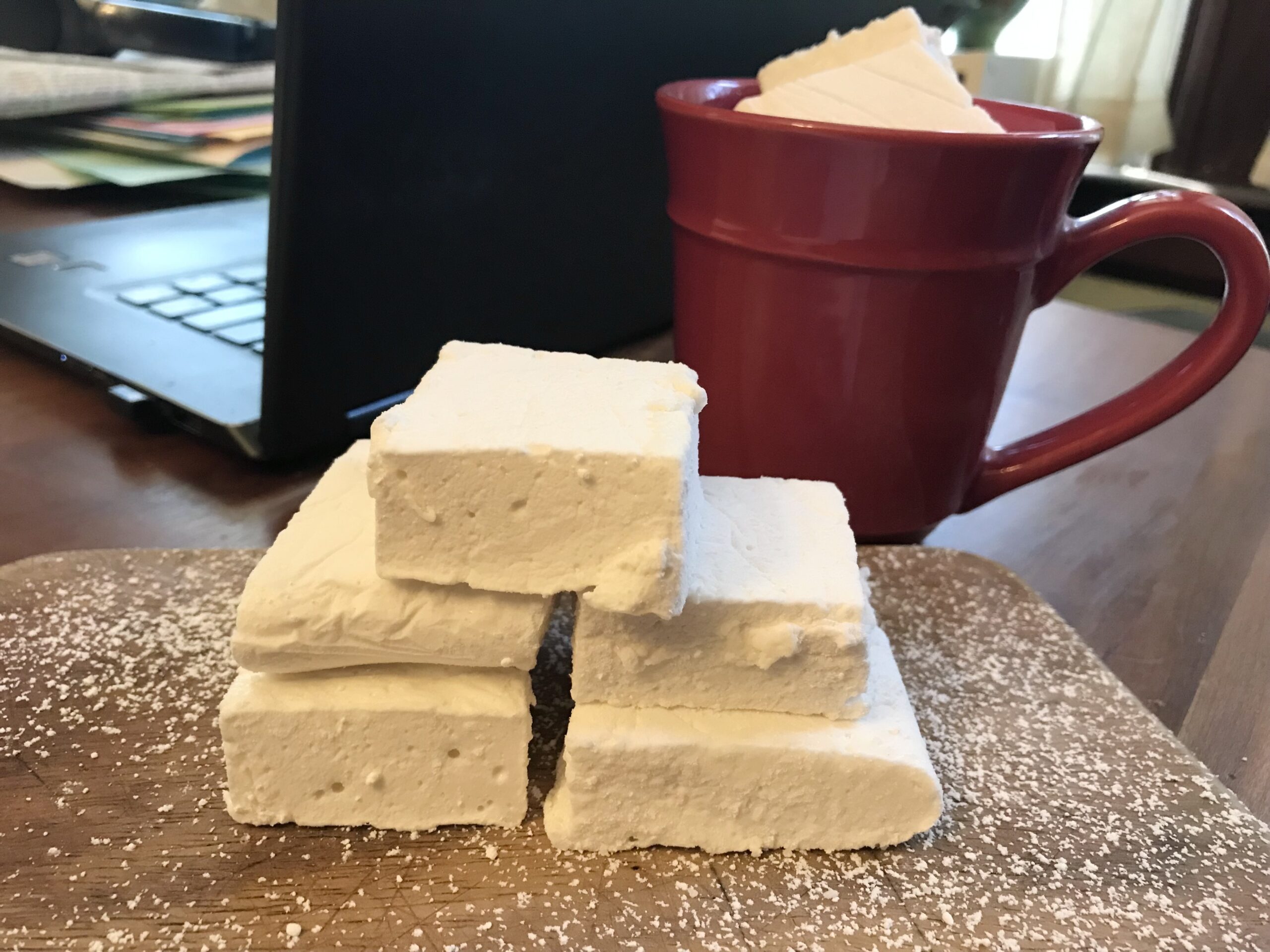 Homemade marshmallows sprinkled with powdered sugar on a cutting board, next to a mug of hot chocolate with a marshmallow in it.