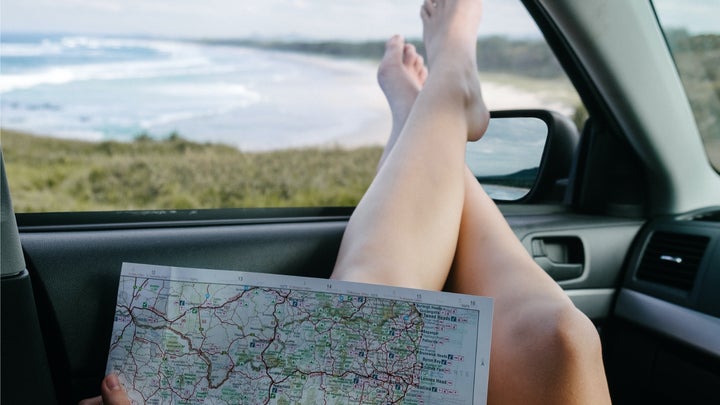 A person sitting in a car with their bare feet out the open window, looking at a road map while overlooking an ocean.