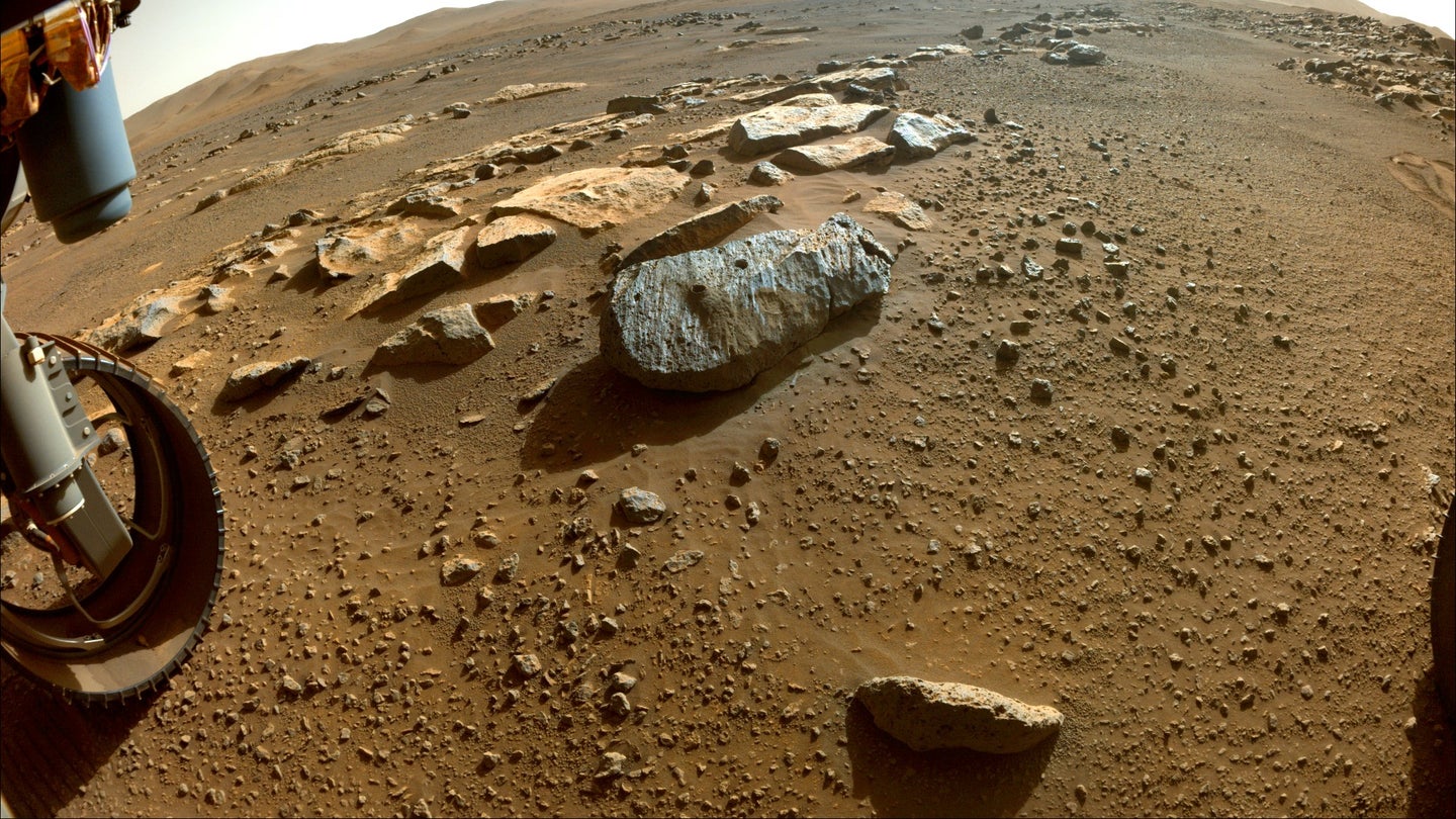 An extremely wide-angle photo of a red, dusty surface, with a grey rock with two holes drilled into it.