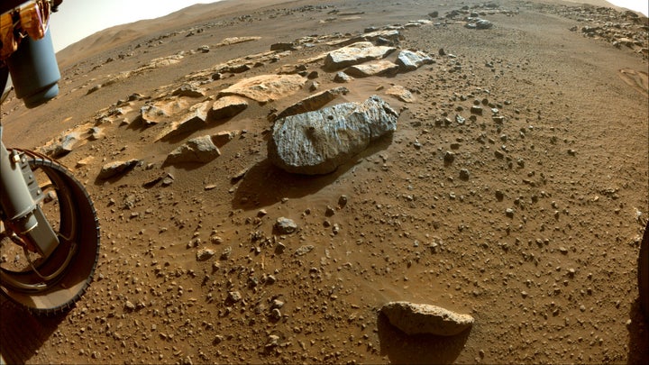An extremely wide-angle photo of a red, dusty surface, with a grey rock with two holes drilled into it.