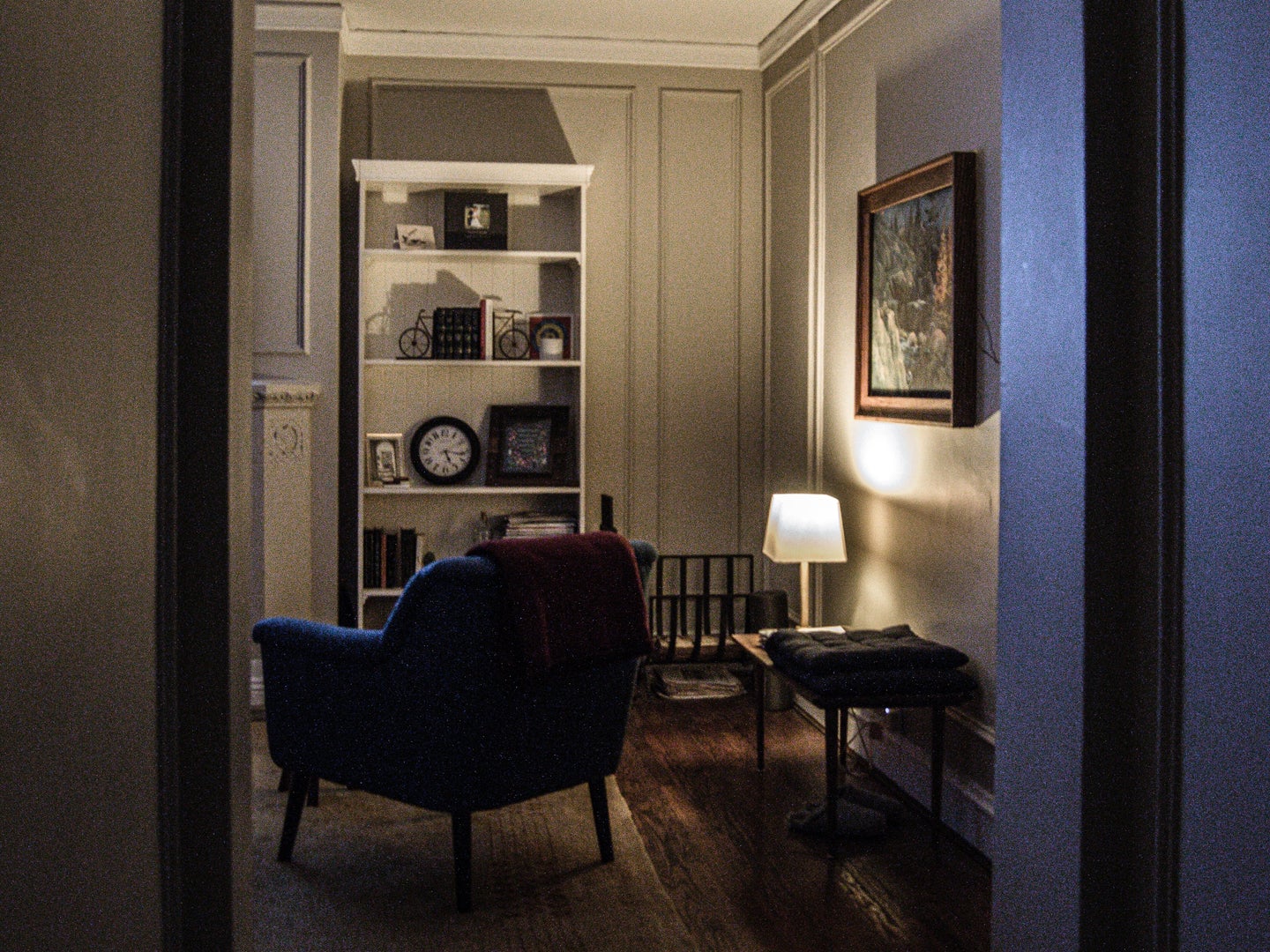 A room with white walls and a hardwood floor with a small lamp in the corner that's lighting the area around a blue armchair.