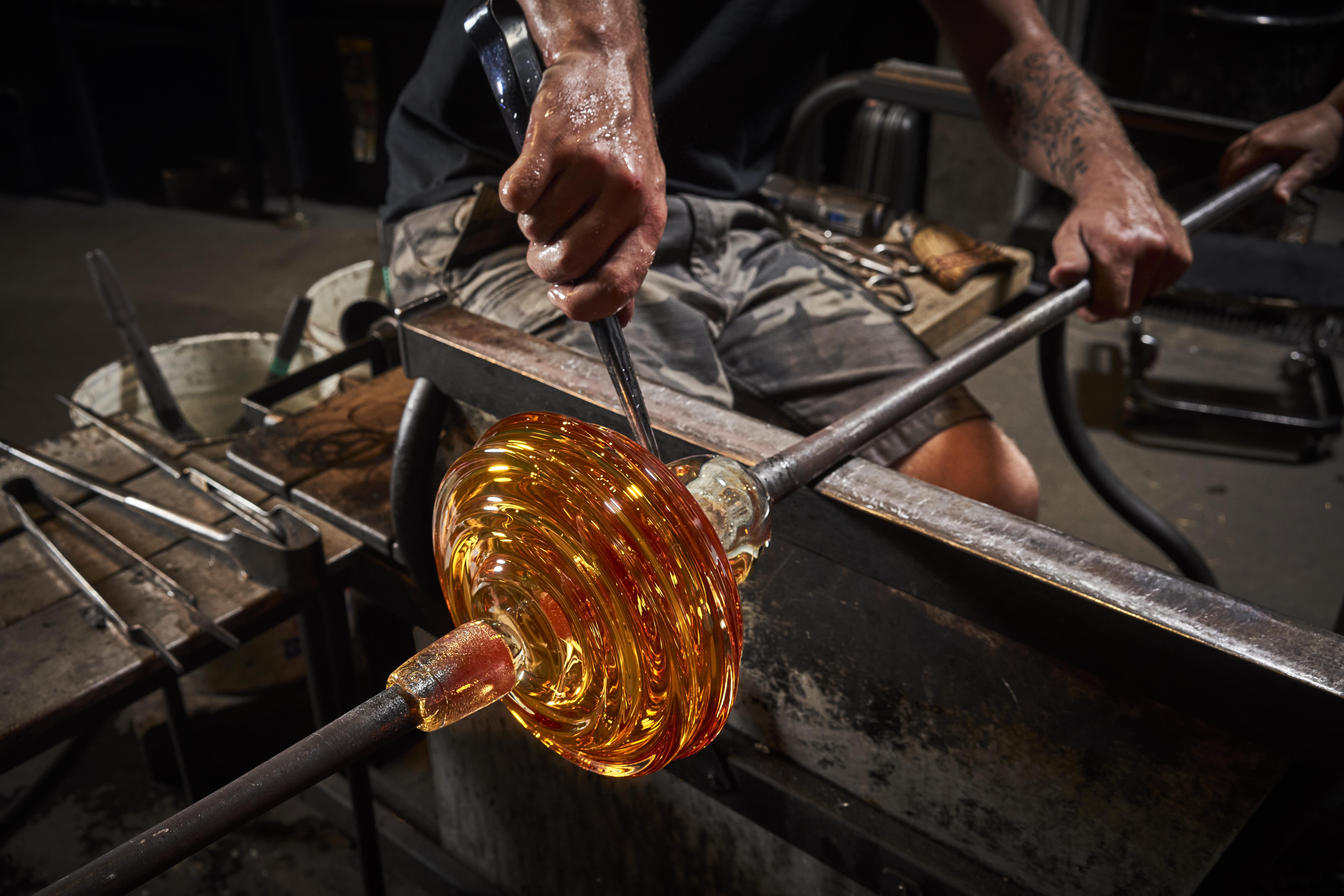 a human's hands hold a metal rod as they manipulate a rounded piece of orange glass