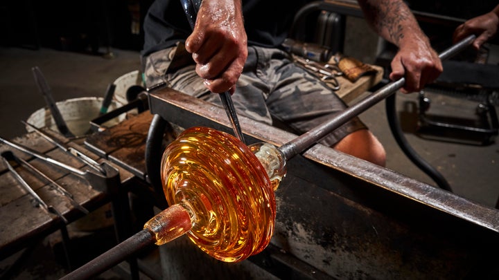 a human's hands hold a metal rod as they manipulate a rounded piece of orange glass