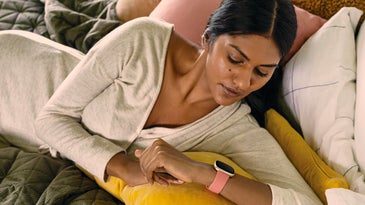 A woman in bed wearing a white long-sleeved shirt, looking at a pink Fitbit on her wrist.