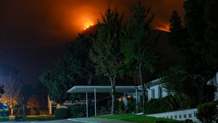 A large home in California being threatened by a wildfire on a distant mountain at night.