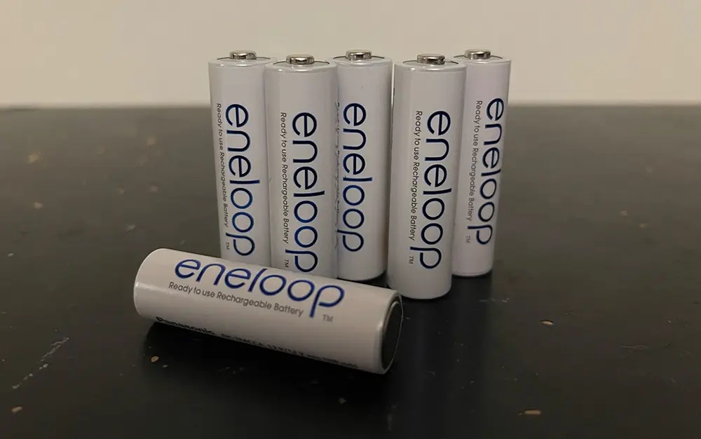 Five AA Panasonic Eneloop batteries standing on a table with a sixth lying down in the foreground in front of them.