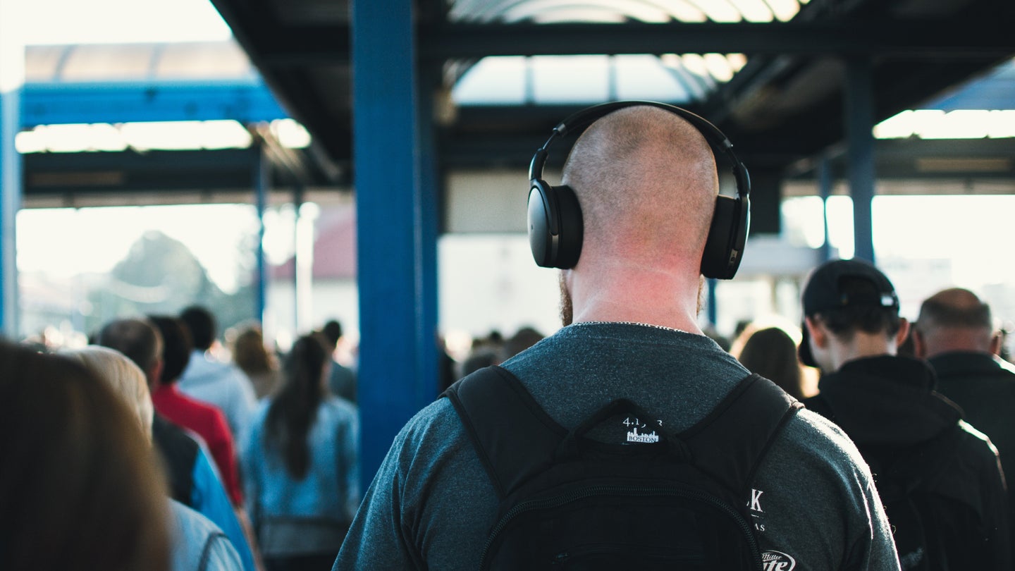A bald man wearing headphones and a backpack in a crowded area, maybe a train station.