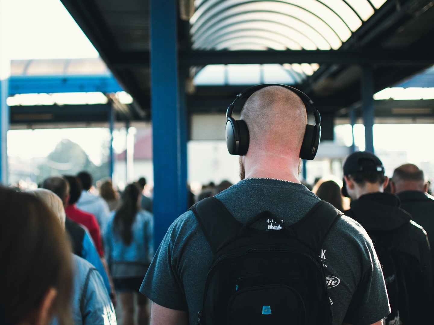 A bald man wearing headphones and a backpack in a crowded area, maybe a train station.