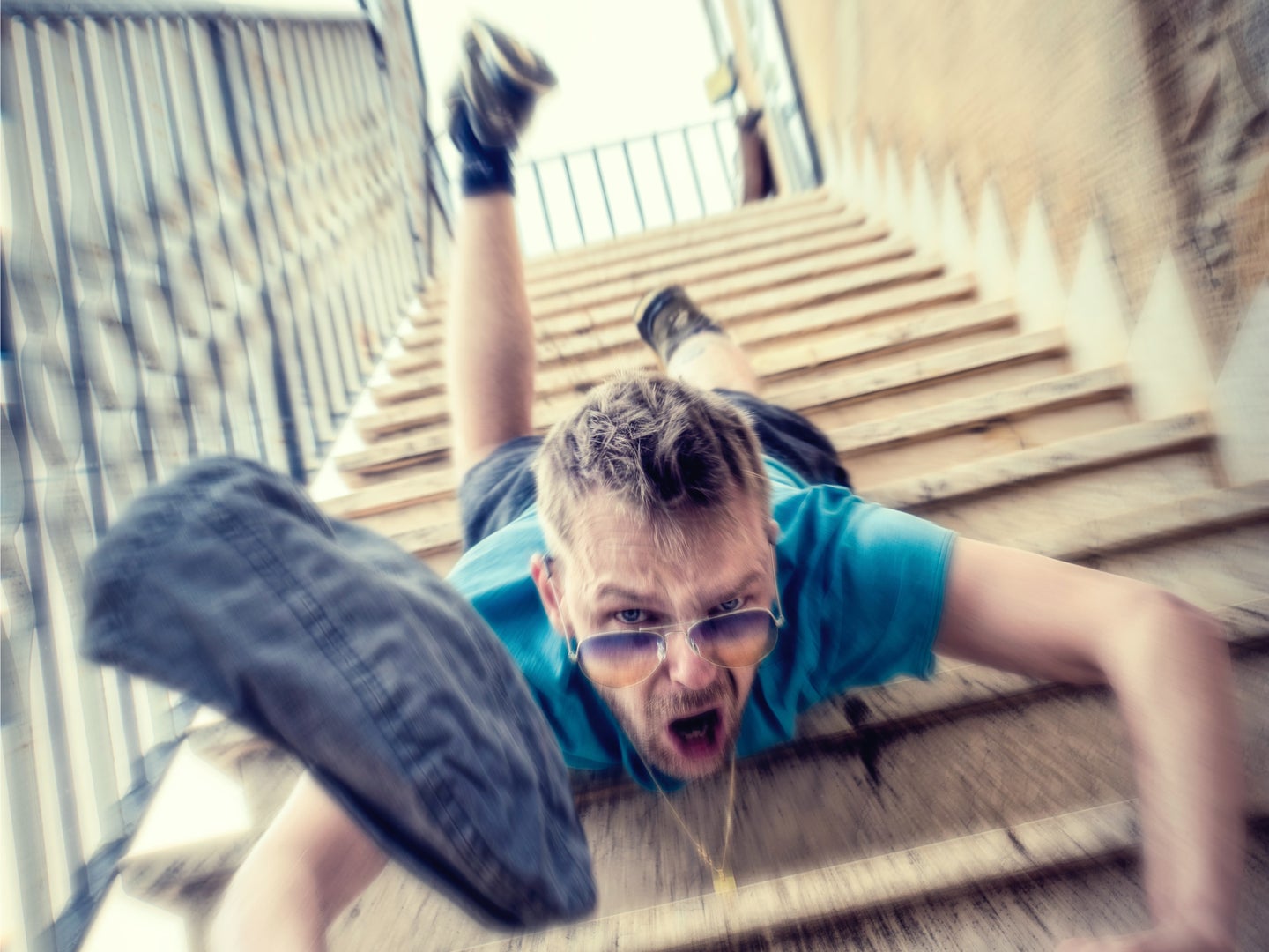 A man wearing sunglasses, a blue t-shirt, and shorts falling face-first down some concrete exterior stairs, while dropping a pair of jeans.