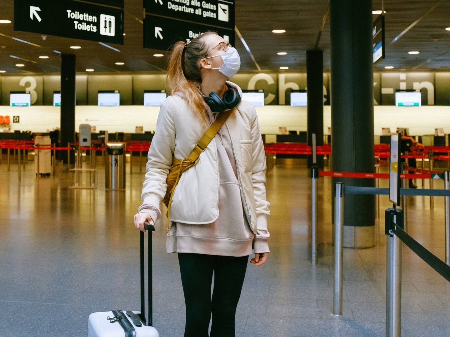 A blonde person wearing a medical face mask and standing in a mostly empty airport while holding onto a white rolling suitcase.