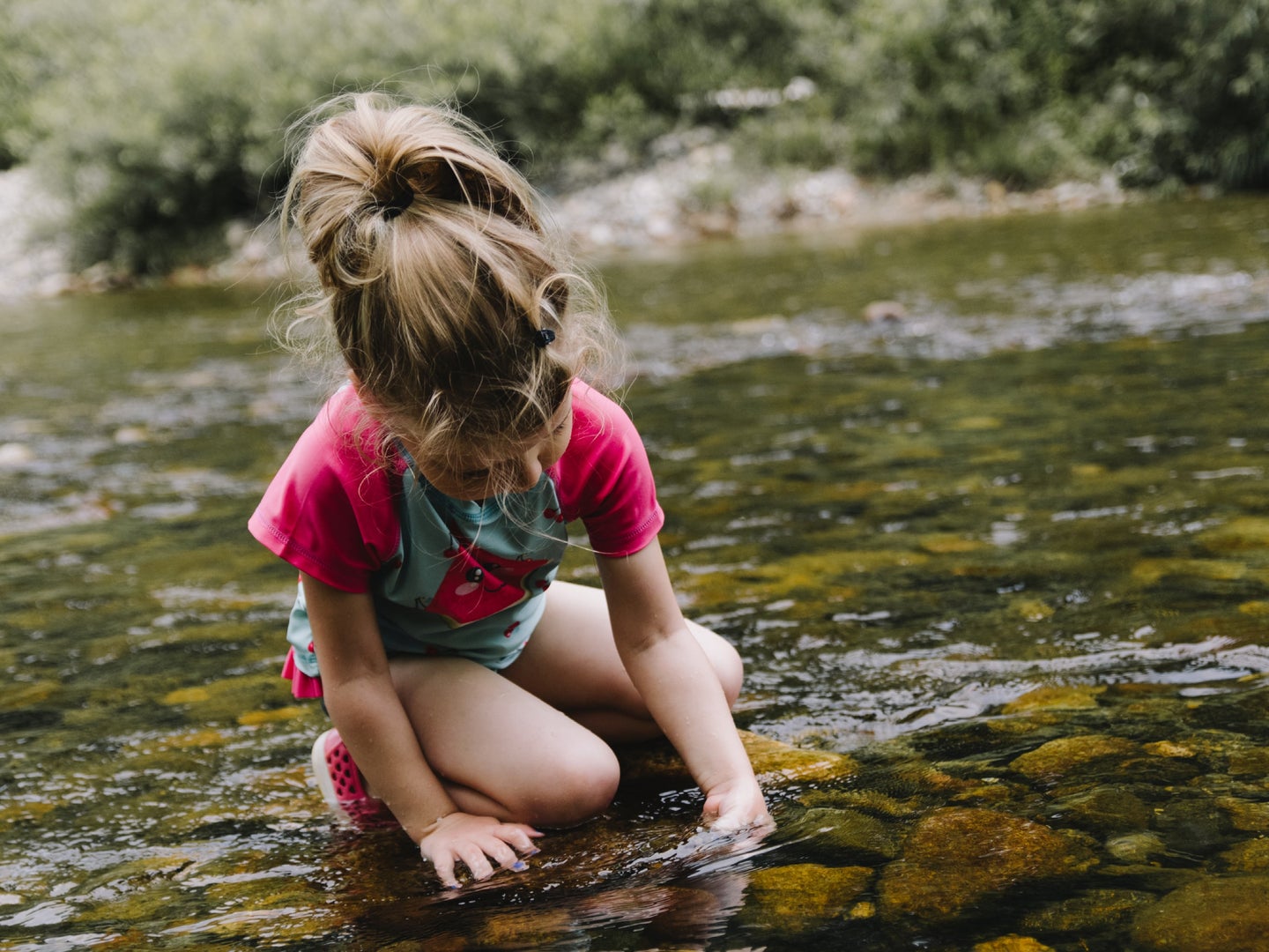 A blonde child wearing pink and green while kneeling in a shallow stream to pick up a rock.