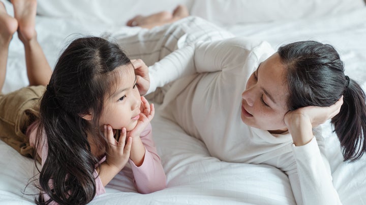 A woman in white and a child wearing a pink shirt and brown pants lying on a white bed, talking.