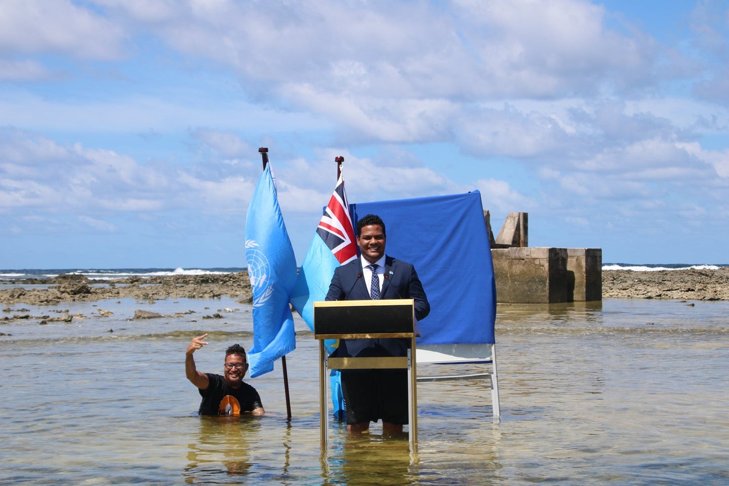 Tuvalu minister Simon Kofe records his COP26 speech in a suit and at a lectern while standing with his flags in the Pacific Ocean