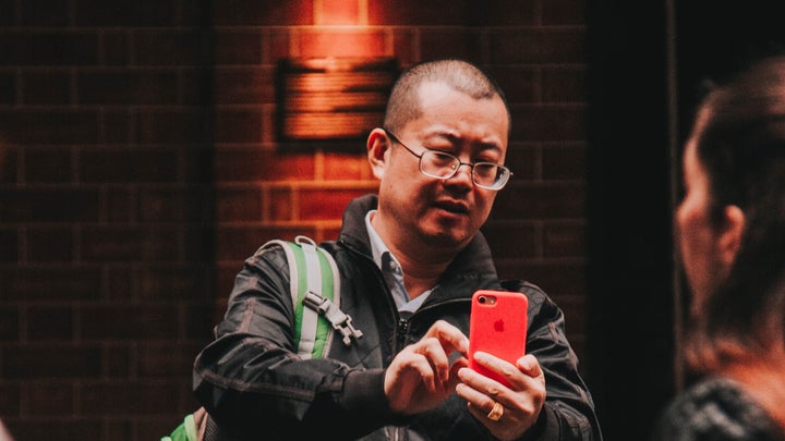 An Asian man with short hair and glasses wearing a black jacket and a green backpack while zooming in on something on the screen of his iPhone.