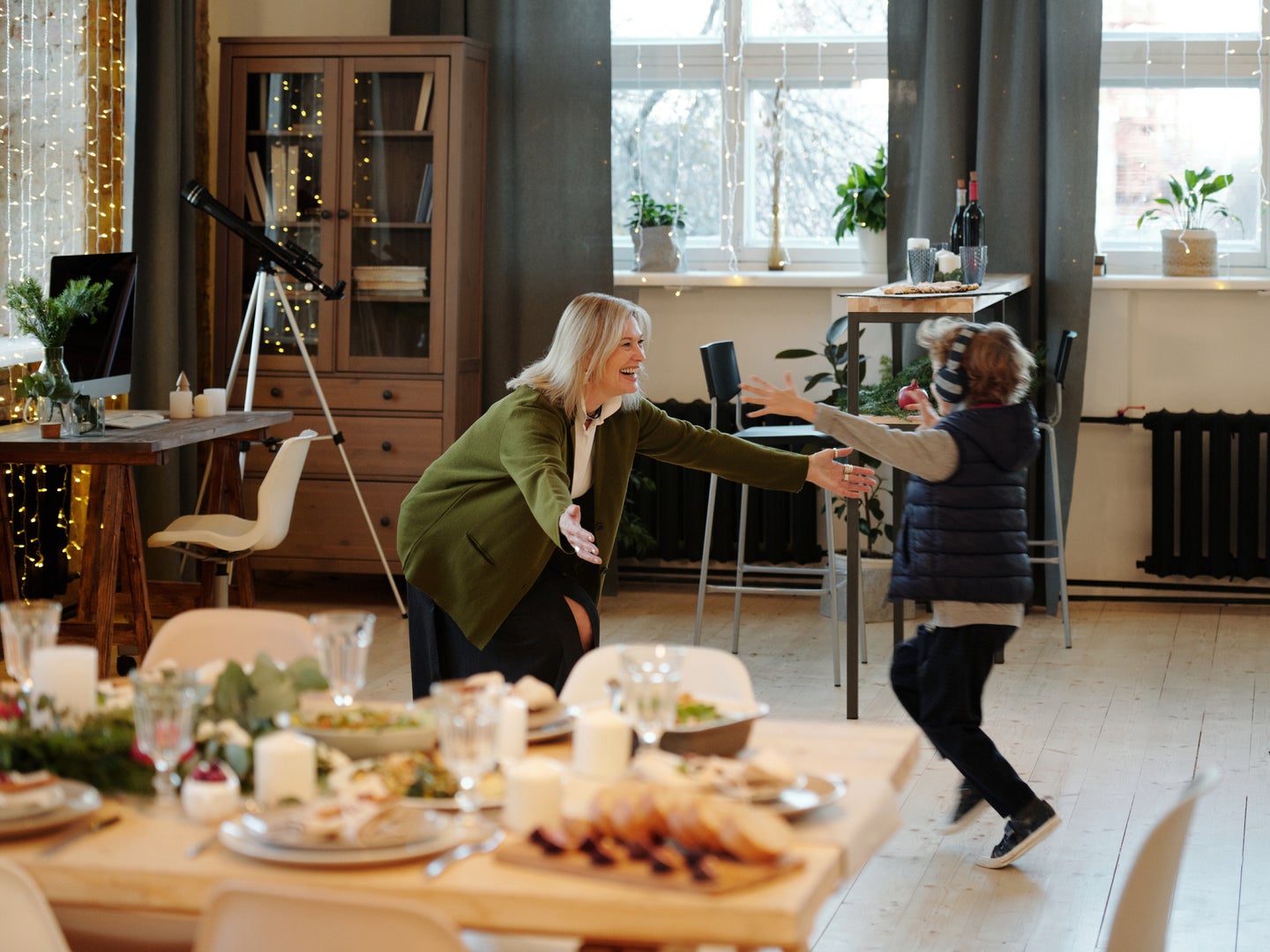 A blonde child running with open arms toward an elderly woman who is ready to hug him, in a room decorated for the holidays with a lot of food on the table.