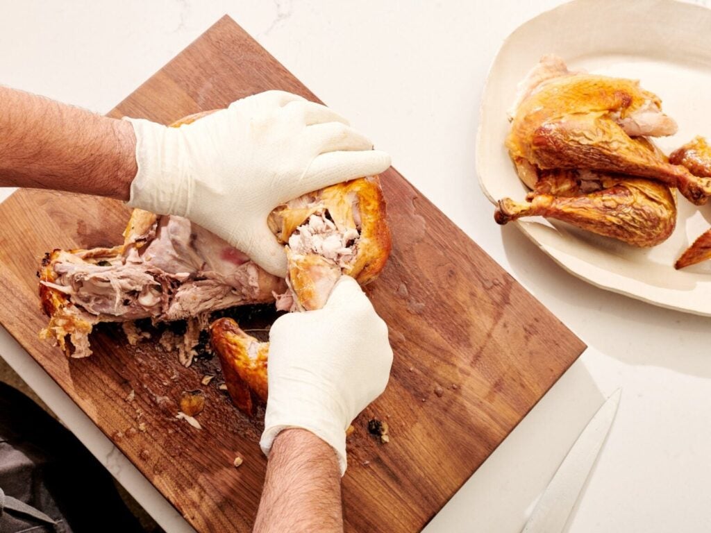 Gloved hands tearing apart a turkey on a wooden cutting board and white plate