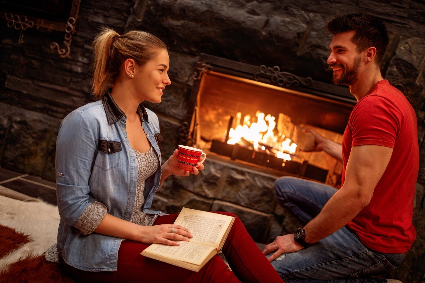 Person with long blond hair holding a book and cup of tea and smiling at a person with short brown hear and a red t-shirt in front of a family fireplace during the holidays