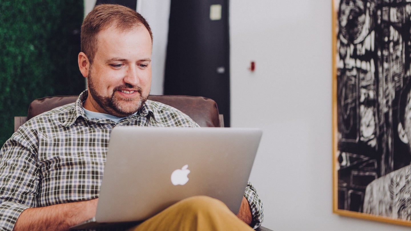 A middle-aged man wearing a green plaid collared shirt with a silver Macbook on his lap. He is grinning as he adjusts some hidden settings to speed up his macOS laptop.