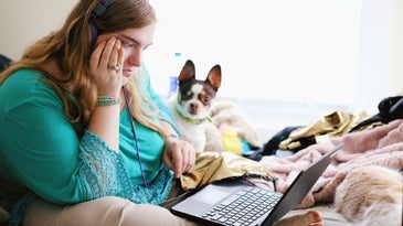 College students in headphones and teal shirt sitting on bed with laptop and small brown and white dog trying to get broadband access