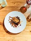 Wild boar ribs, sunchoke chips, and Japanese barberry demiglace on a white plate and wooden table with a glass of red wine